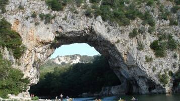 A natural rock arch formation with a river flowing through it, surrounded by greenery and a clear sky.