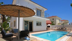 A pool area with lounge chairs, a thatched umbrella, and a white building with a balcony. - Villa Amarilia - 100m from Pernera Beach (Pernera)