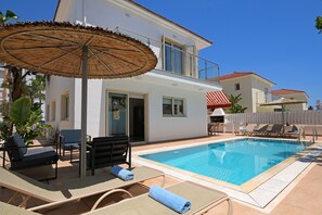 Un espace piscine avec des chaises longues, un parasol en chaume et un bâtiment blanc doté d’un balcon.