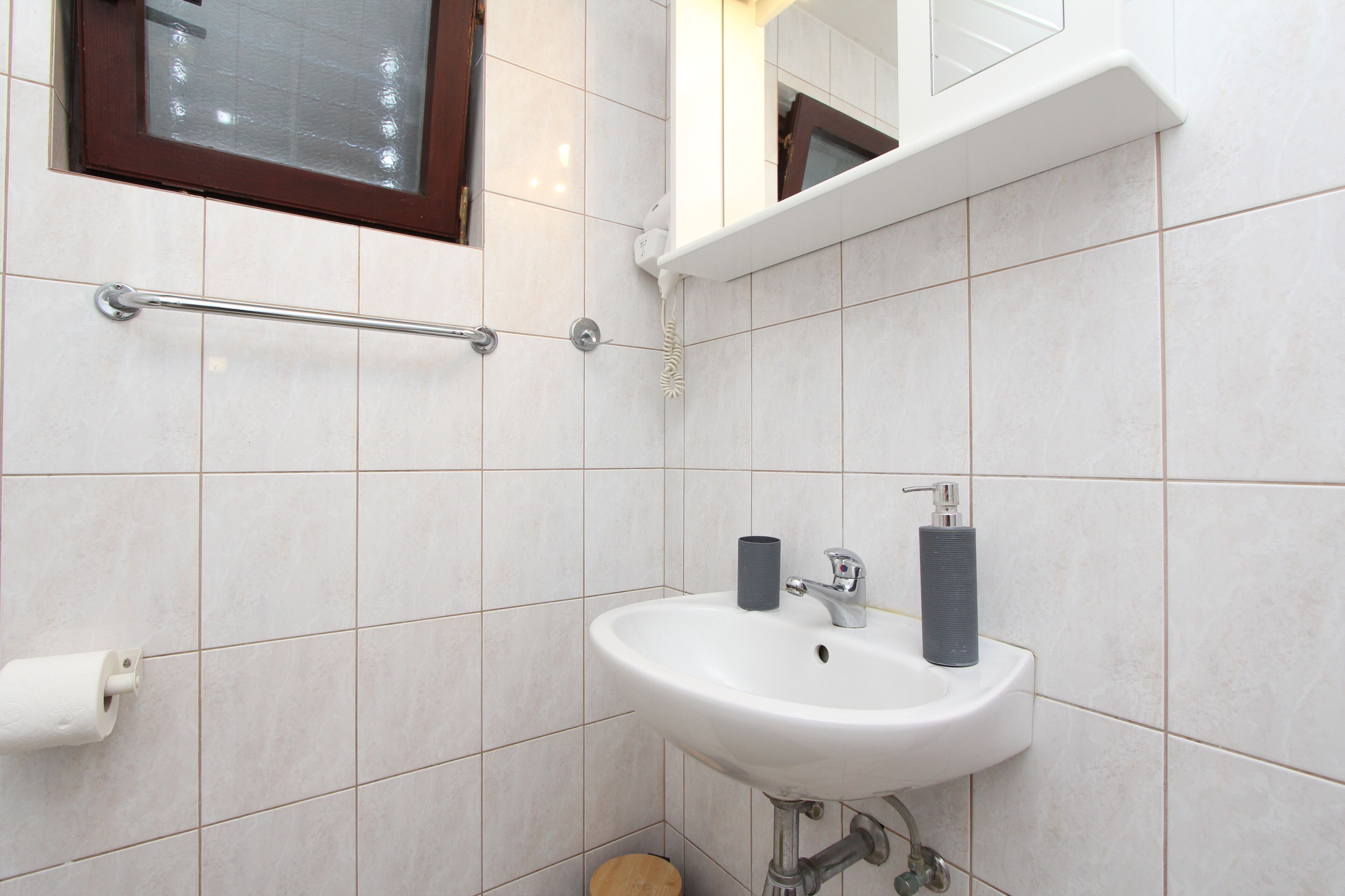 A bathroom with white tiles, a white sink with two soap dispensers, a handrail, and a toilet paper holder.