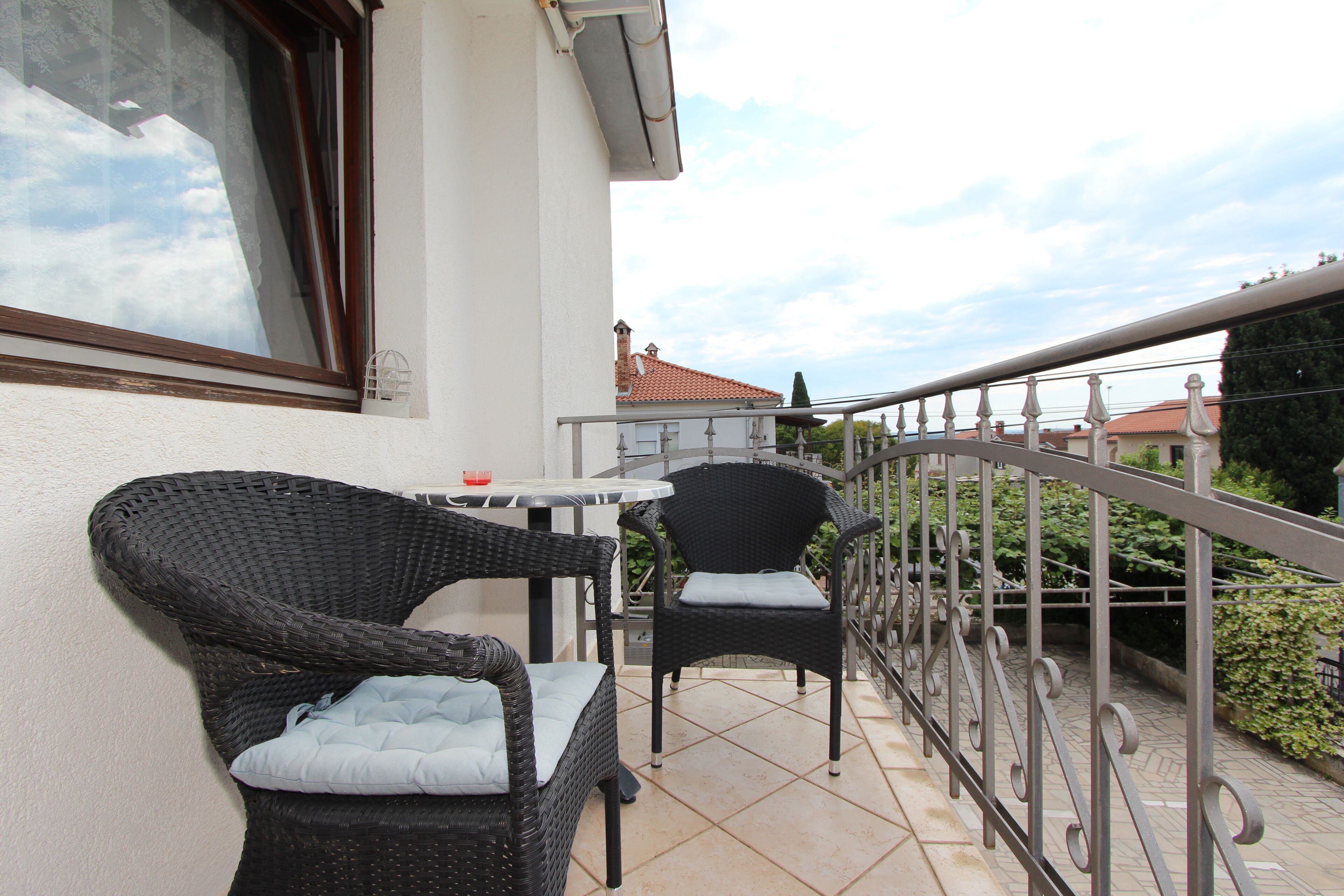 A balcony with wicker chairs and a small table, a metal railing, and a building with a red roof in the background.