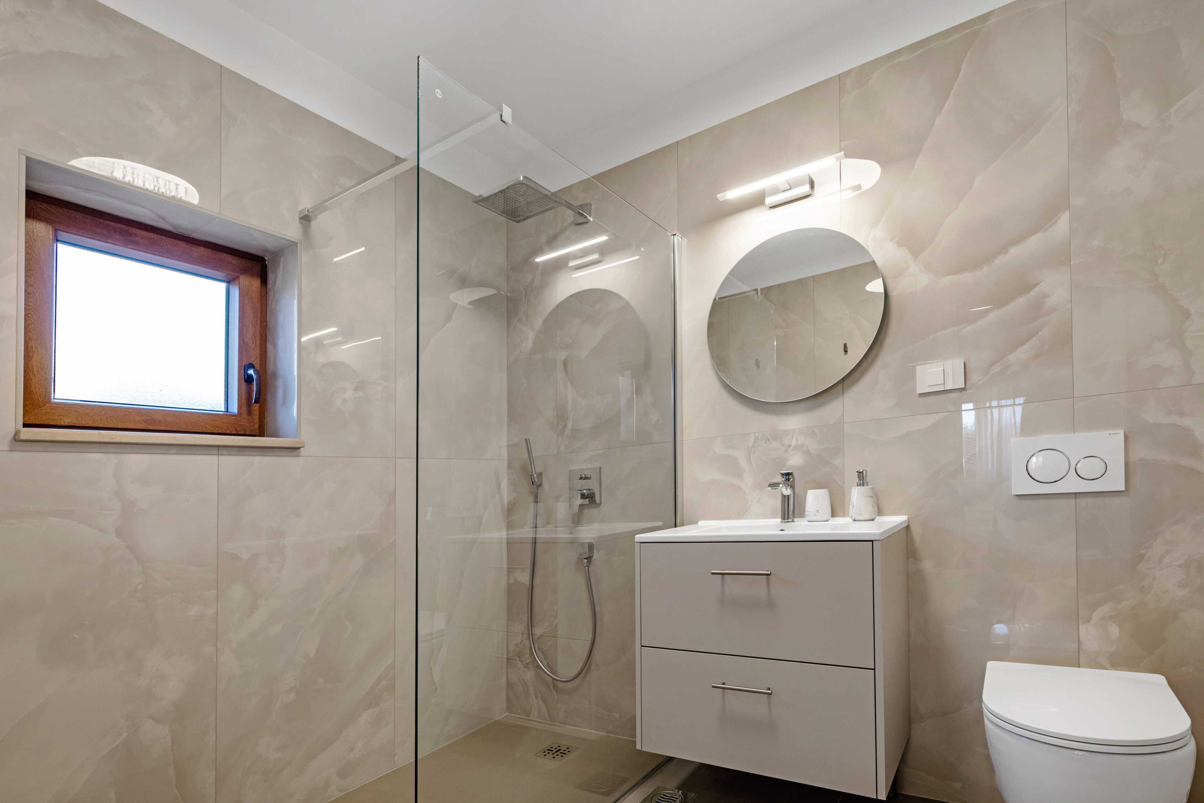 Un bagno moderno con cabina doccia in vetro, uno specchio rotondo, un lavabo bianco e un water.