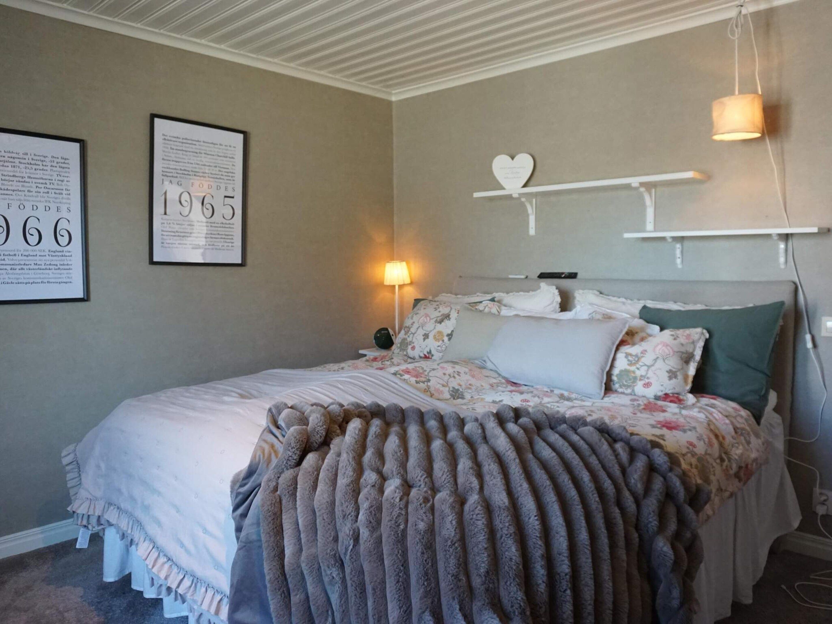 A bedroom with a bed, a nightstand, a lamp, a wall-mounted shelf with a heart-shaped decoration, and two framed documents on the wall.