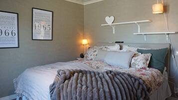 A bedroom with a bed, a nightstand, a lamp, a wall-mounted shelf with a heart-shaped decoration, and two framed documents on the wall.
