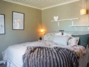 A bedroom with a bed, a wall clock, a framed document, a heart-shaped decoration, and a lamp.