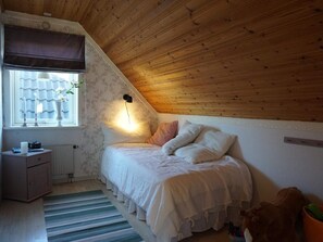 Ein Schlafzimmer mit schrägen Wänden, einem Bett mit weißer Bettwäsche, einem Nachttisch mit Lampe und einem Fenster mit Vorhängen.