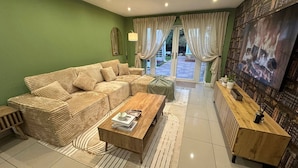 A living room with a sectional sofa, a wooden coffee table, a TV, and a view through glass doors to an outdoor area.