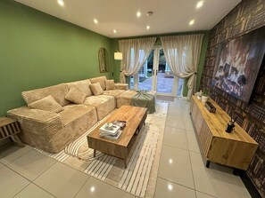 A living room with a sectional sofa, a wooden coffee table, a TV, and a view through glass doors to an outdoor area.