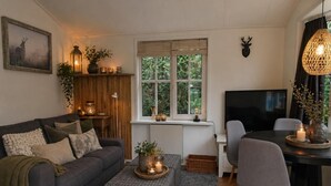 A cozy living room with a grey sofa, a small table with candles, a TV, and a window with a view of greenery. - Vechtdal Chalet de Berk (Rheezerveen)
