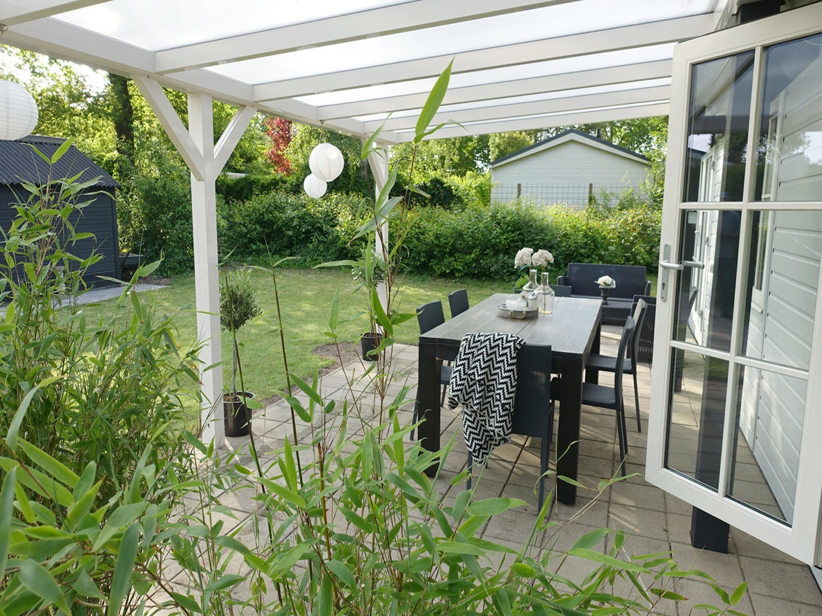 A covered patio with a table and chairs, surrounded by greenery and a house in the background.