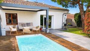 Ein rechteckiger Swimmingpool mit Holzterrasse, eine überdachte Sitzgelegenheit mit Stühlen und Tisch sowie ein Haus mit Ziegeldach und Garten.
