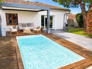 Ein rechteckiger Swimmingpool mit Holzterrasse, eine überdachte Sitzgelegenheit mit Stühlen und Tisch sowie ein Haus mit Ziegeldach und Garten.