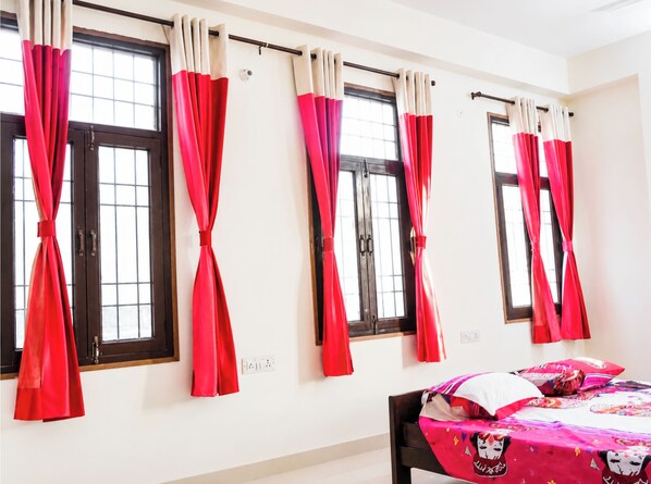 Una habitación con cuatro ventanas, cada una con cortinas rojas atadas, una cama con colcha de colores y una pared blanca.
