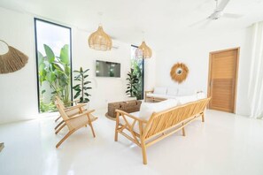 Ein heller Raum mit einer Couch, zwei Sesseln, einem Fernseher und großen Fenstern.