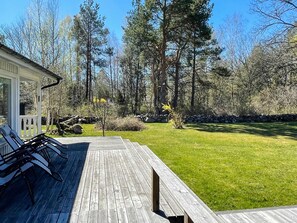 Eine Holzterrasse mit Gartenmöbeln, eine Steinsäule und eine Rasenfläche mit Bäumen.