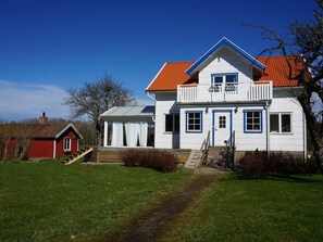 A two-story house with a red roof, white walls, and blue trim. There is a small red shed to the left and a staircase leading to the front door.
