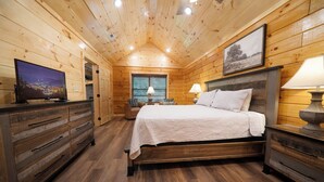 Ein Schlafzimmer einer Holzhütte mit Bett, Kommode, Fernseher und einem Fenster mit Jalousien.