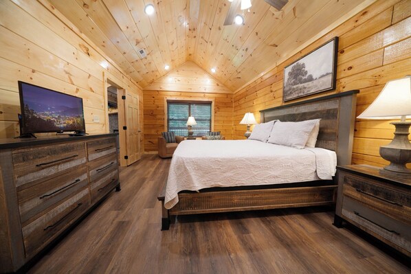 A wooden cabin bedroom with a bed, dresser, TV, and a window with blinds. - Enchanted Pines: Find Your Magic in the Mountains (Pigeon Forge)