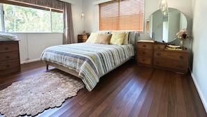 Un dormitorio con cama, cómoda de madera, mesita de noche, ventana con persianas, aire acondicionado y un espejo redondo.