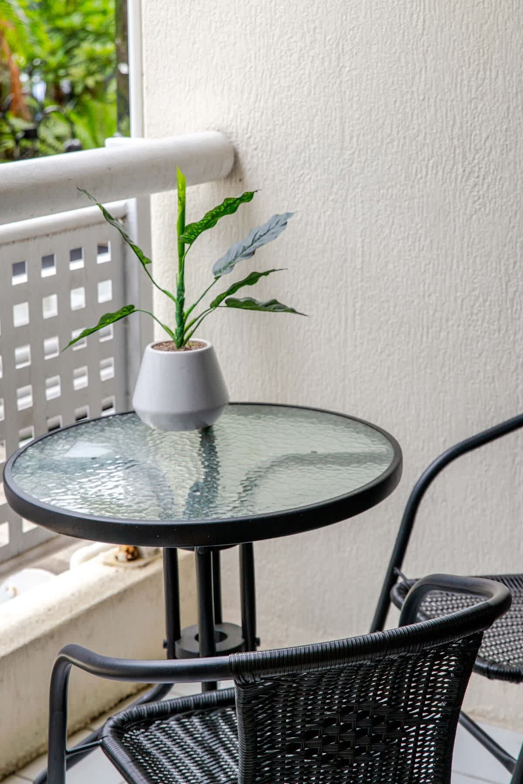 Een ronde glazen tafel met een witte vaas met een plant, zwarte rieten stoelen en een witte tralieschutting.