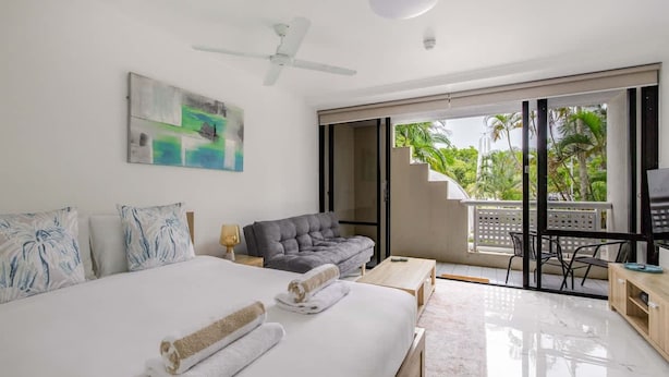 Habitación de hotel con cama, sofá gris, escritorio y balcón con vistas a palmeras.