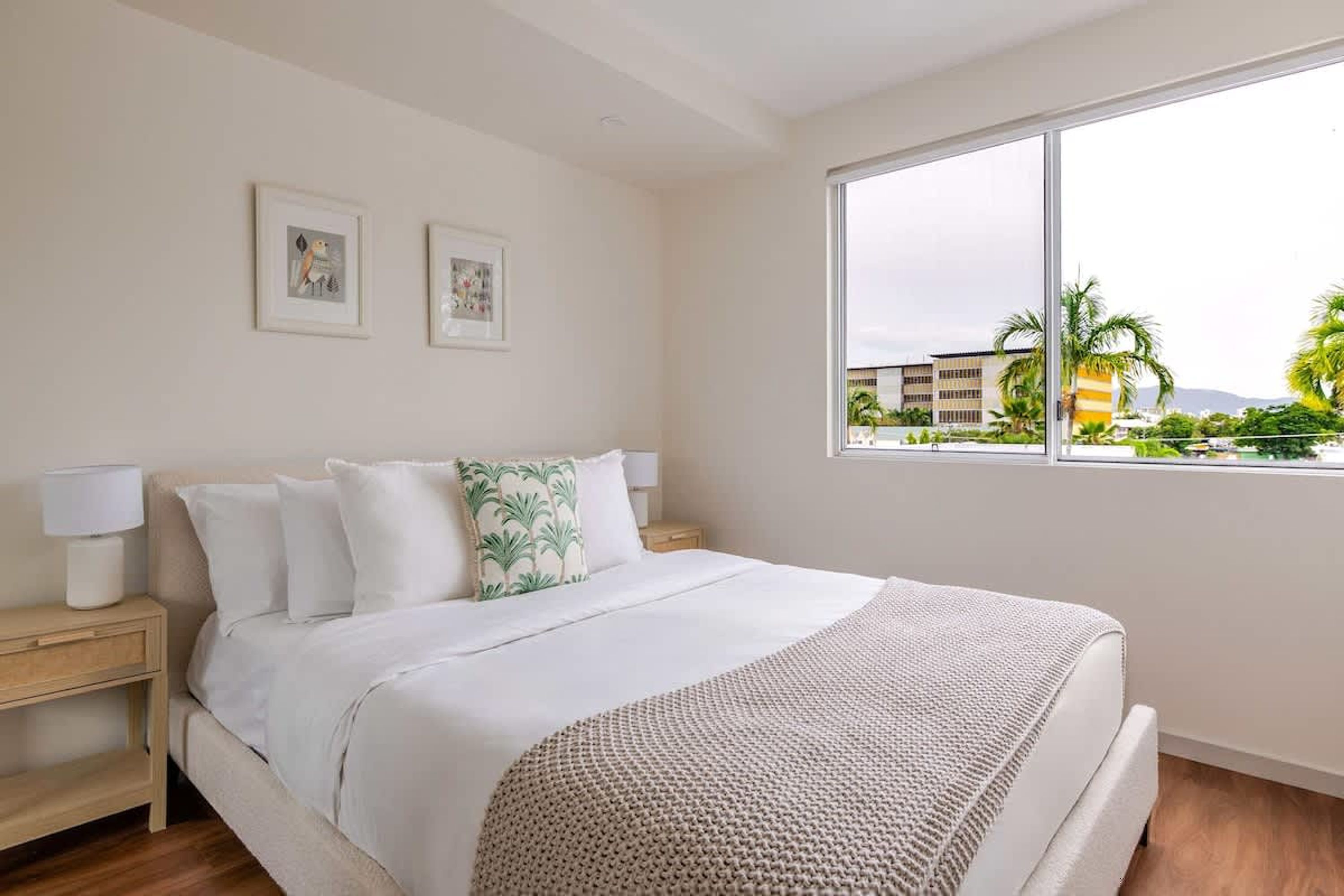 A bedroom with a bed, bedside tables, a window with a view of palm trees and buildings.