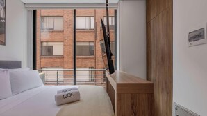 A modern hotel room with a large bed, a wooden nightstand, a window with a view of a brick building, and a mounted rifle. - NOK Cozy 1BR Apartment in Prime Bogota Location (Bogotá)