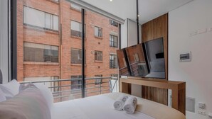 A modern hotel room with a large bed, a TV on a wooden stand, and a view of a brick building through large windows. - NOK Cozy 1BR Apartment in Prime Bogota Location (Bogotá)