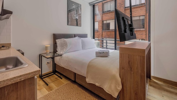 A modern hotel room with a bed, bedside table, lamp, and a large window with a view of a brick building. - NOK Cozy 1BR Apartment in Prime Bogota Location (Bogotá)