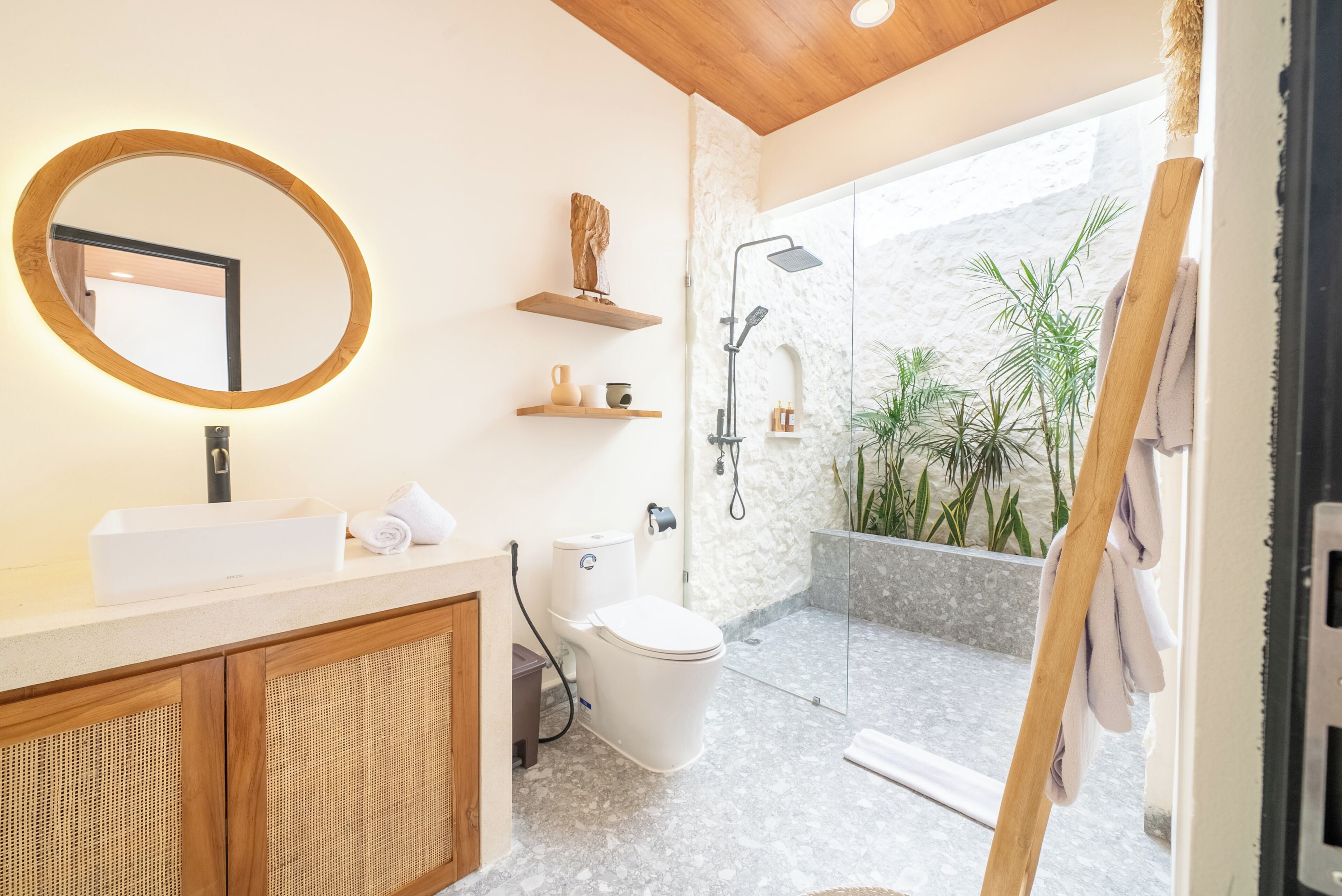 A modern bathroom with a wooden vanity, a round mirror, a toilet, a shower area, and a view of the beach.