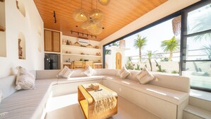Een moderne woonkamer met een grote witte bank, een houten salontafel en uitzicht op palmbomen door grote ramen.