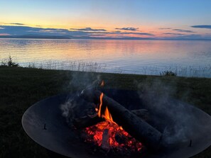 A campfire with smoke rising, surrounded by grass and overlooking a calm body of water with a sunset sky. - Water's Edge Cottage - New 2026! (Castine)