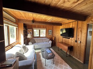 A wooden-paneled room with a sofa, a coffee table, a TV, and a bookshelf. - Water's Edge Cottage - New 2026! (Castine)
