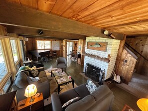 A cozy cabin interior with a fireplace, wooden walls, a dining area, and a piano. - Water's Edge Cottage - New 2026! (Castine)