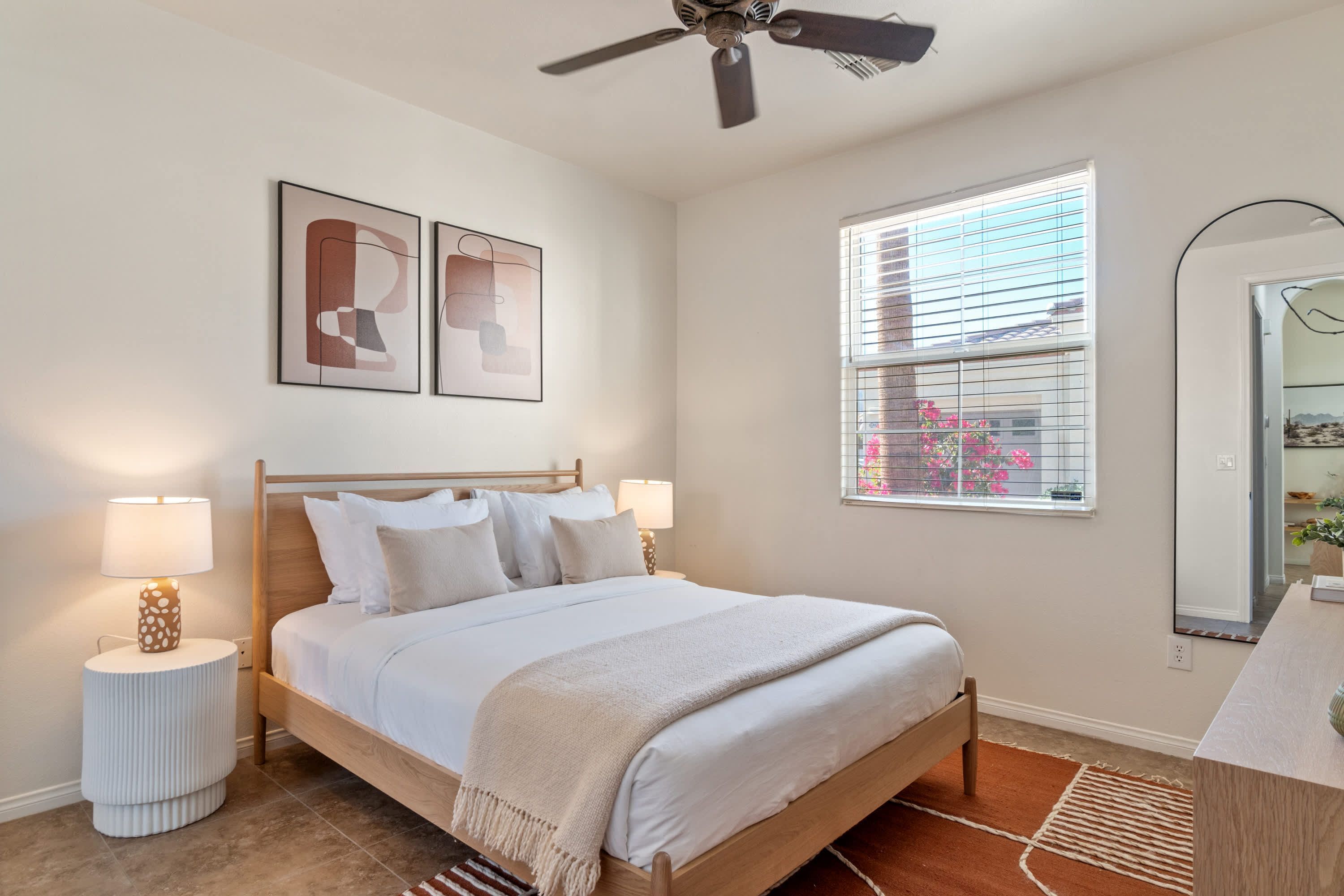 A bedroom with a bed, bedside lamps, a ceiling fan, a window with blinds, and a mirror.