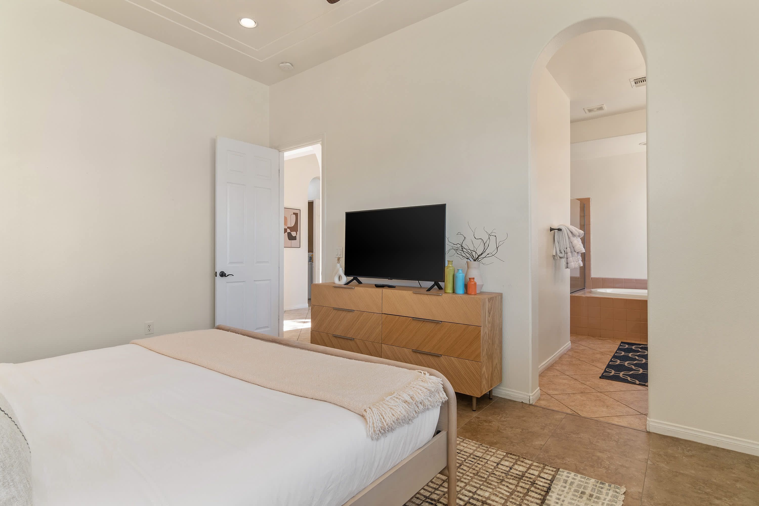 A bedroom with a bed, a TV on a dresser, a bathroom visible through an archway, and a tiled floor.