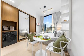 A modern kitchen with a washing machine, a dining area with a table set for two, and a large window with a city view. - Free Housekeeping Visits During Your Stay - Stayshort - Studio in Jumeirah Village Circle for 2 With Balcony (Dubai)