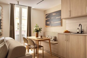 Ein modernes Wohnzimmer mit einer Couch, einem Esstisch mit Stühlen, einer Küchenzeile inklusive Schränken und Spüle sowie einem großen Fenster mit Vorhängen.