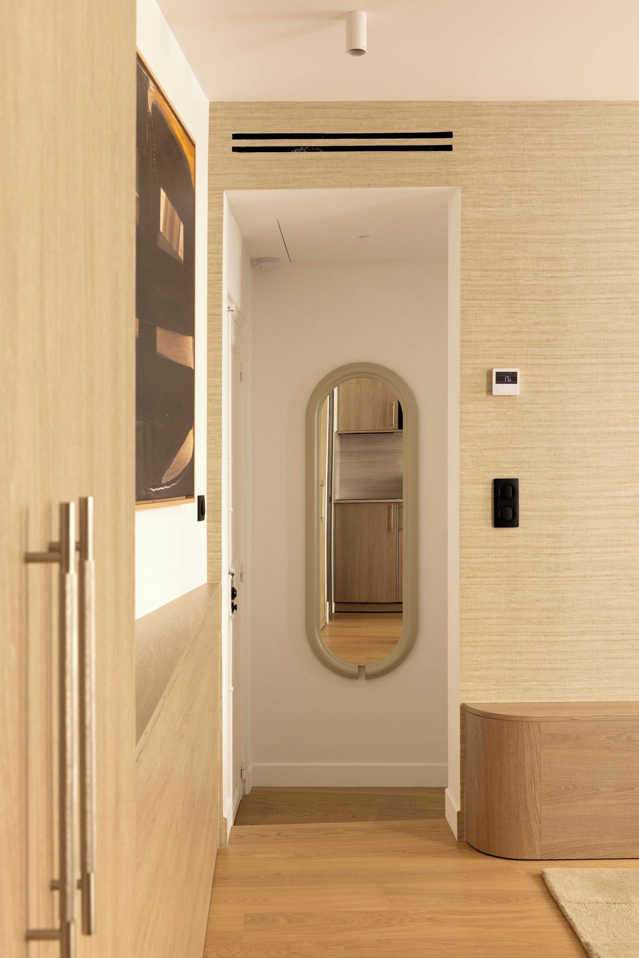A hallway with a wooden floor, a door with a circular mirror, and a wall-mounted light fixture.