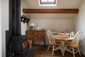 A cozy room with a wood-burning stove, a dining table with chairs, and a window above the stove. - The Damson Barn - Luxury Cottages (Brown Edge)