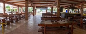 Un espacio al aire libre cubierto con bancos y mesas de madera, pilares de ladrillo y vistas al exterior.