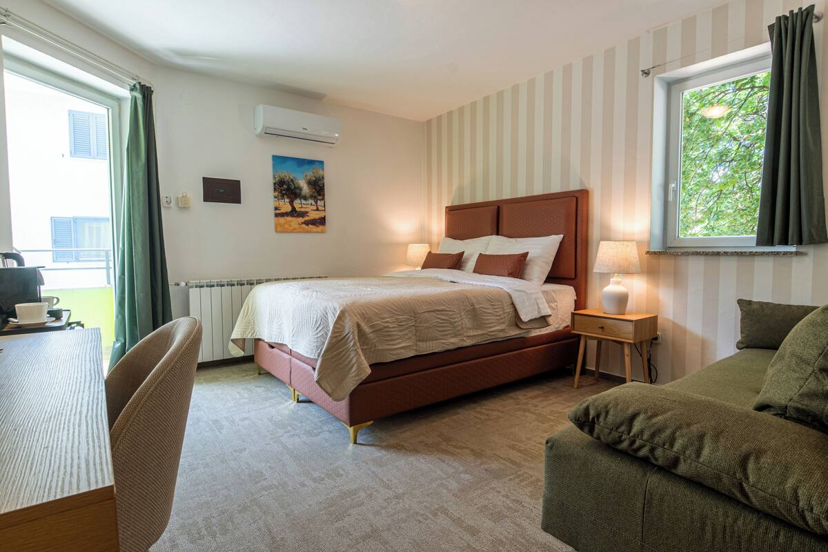 Ett hotellrum med en säng, ett skrivbord, en stol, ett nattduksbord med en lampa och ett fönster med gardiner.