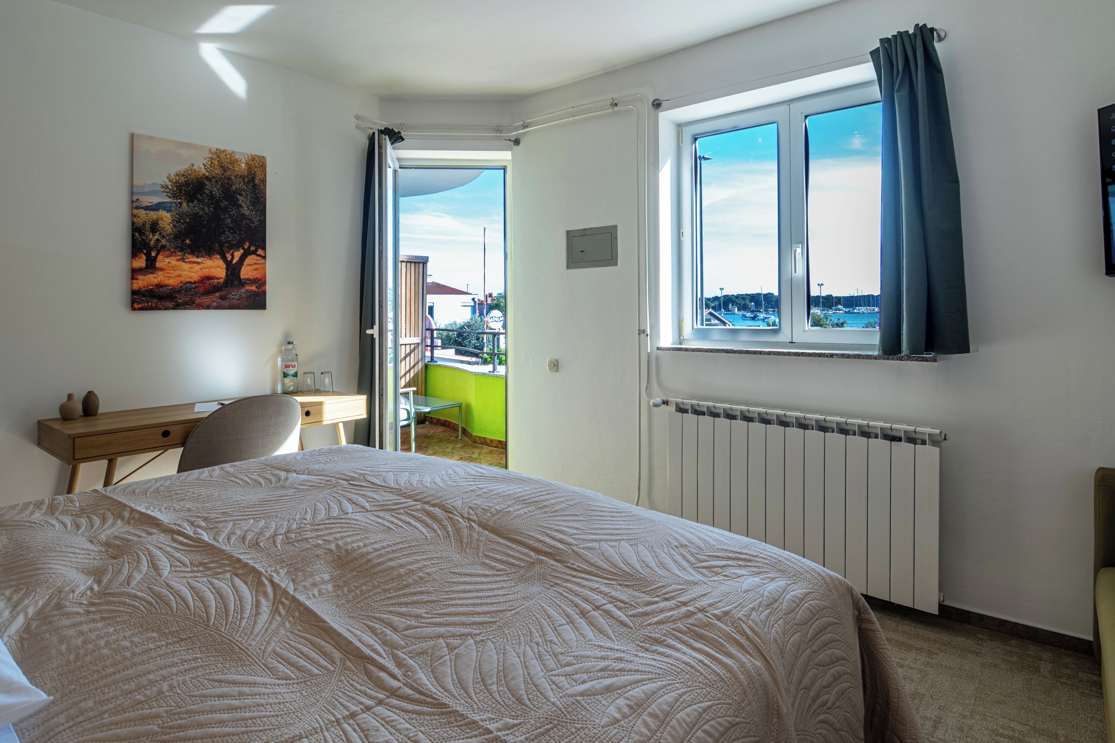A bedroom with a bed, a desk, a chair, a radiator, a window with curtains, and a painting on the wall.