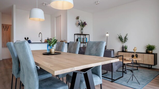 A modern dining area with a wooden table, grey upholstered chairs, and pendant lighting. - Apartment in Breskens Near the Beach (Breskens)