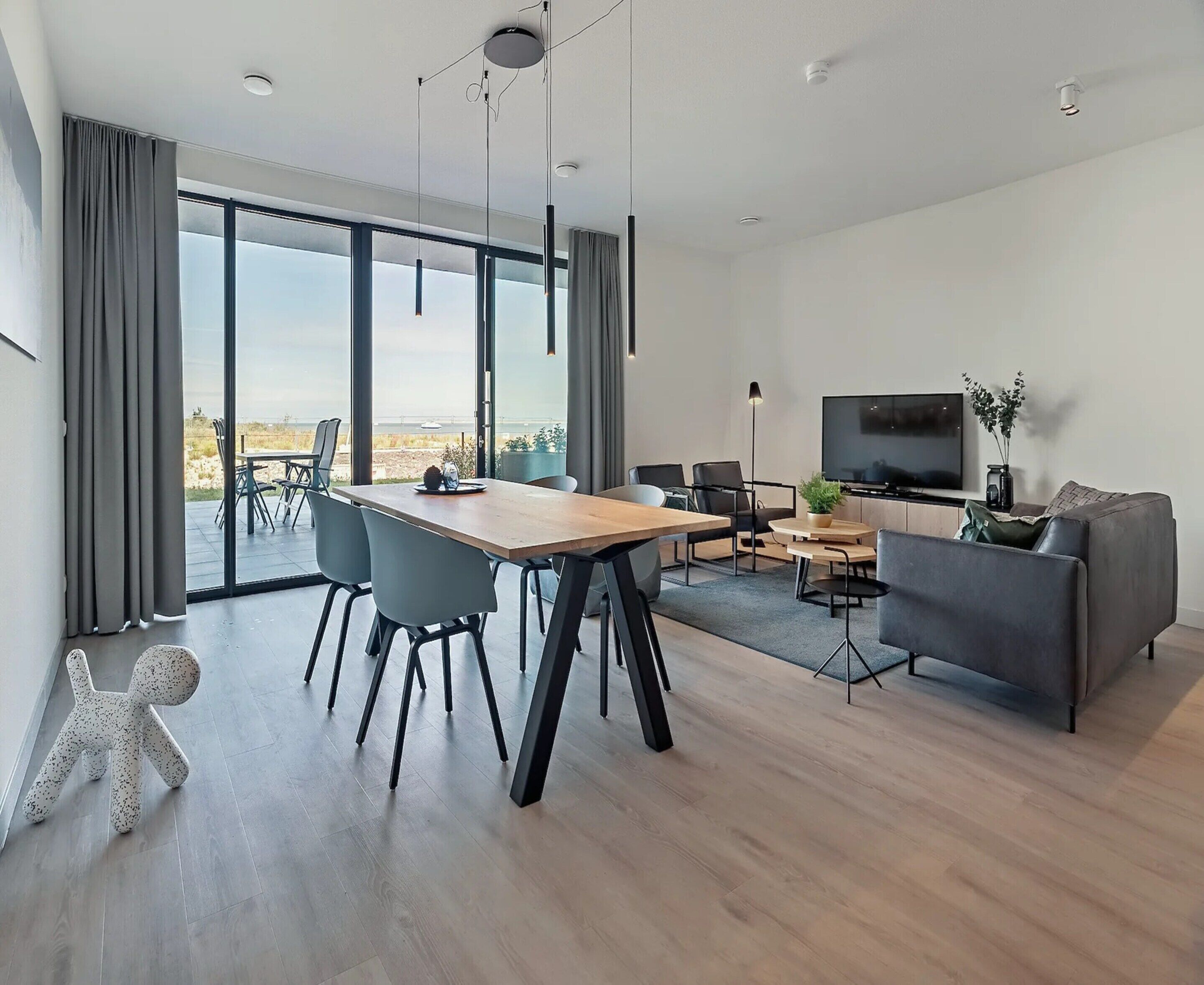 A modern living room with a dining area, large windows, a flat-screen TV, and a grey sofa.