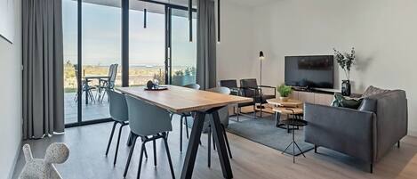 A modern living room with a dining area, large windows, a flat-screen TV, and a grey sofa.