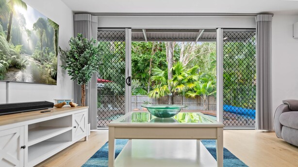 Uma sala de estar com uma mesa de vidro, um armário branco, um tapete azul e vista para um jardim através de uma porta de correr.