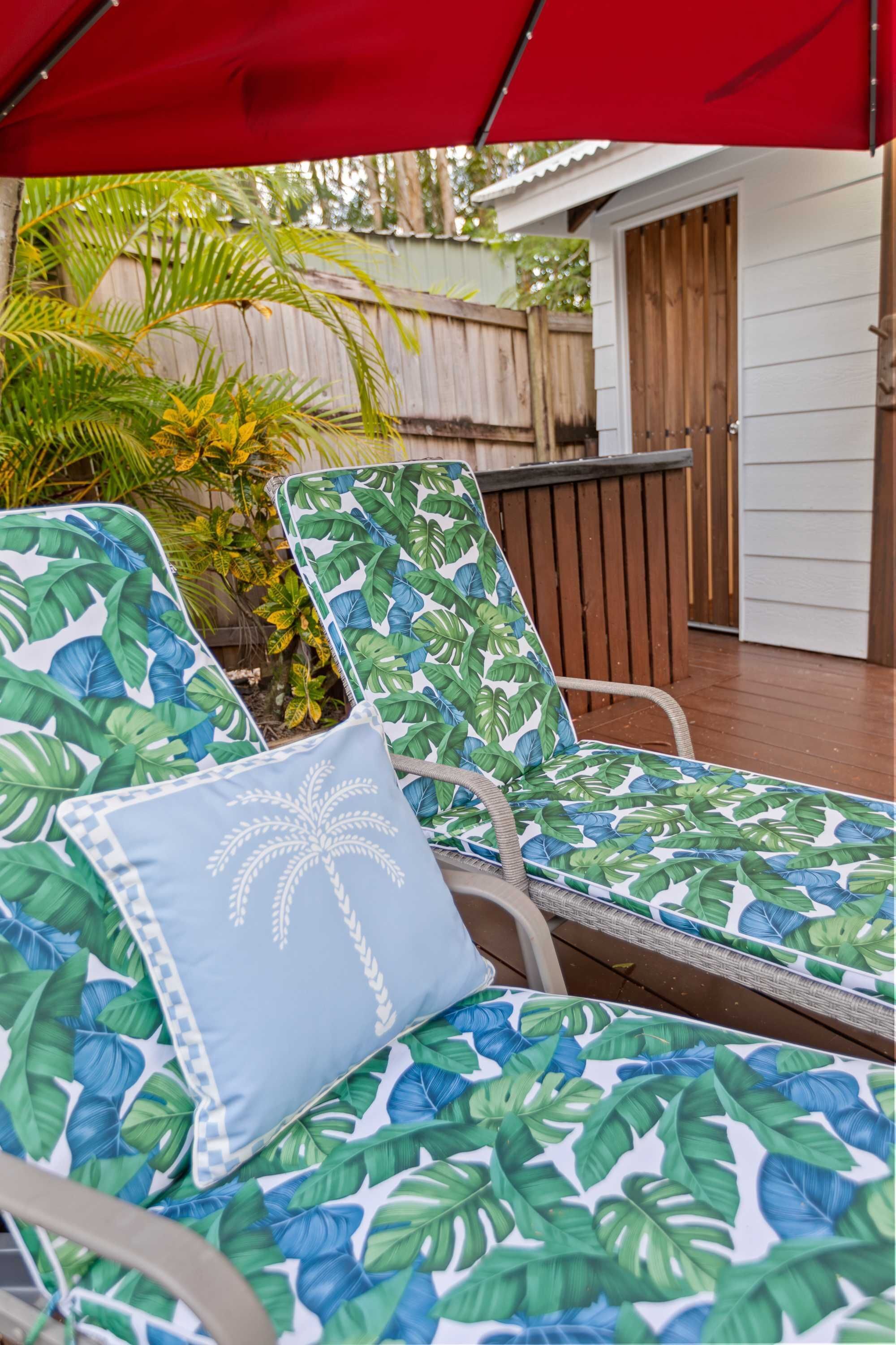 A patio with two lounge chairs, one with a palm tree cushion, and a red umbrella.