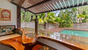 Cozinha com bancada de madeira, pia de aço inoxidável e vista para uma piscina e vegetação tropical.