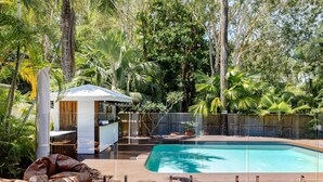Piscina cercada por vegetação exuberante, um deck de madeira e uma pequena estrutura branca com telhado.